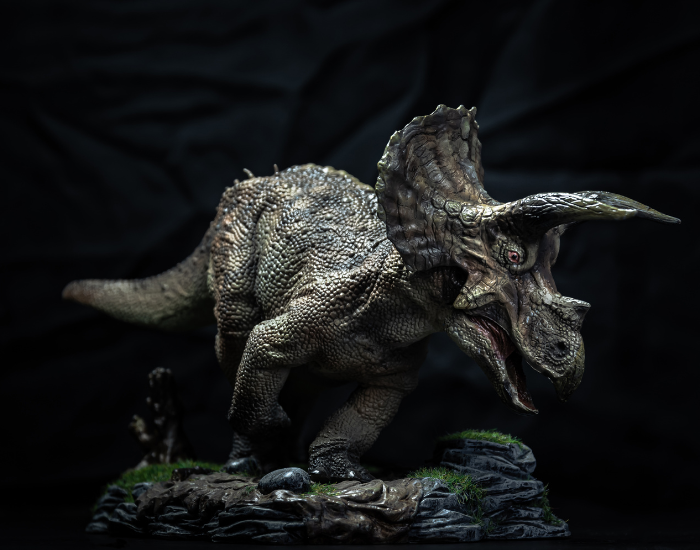 A highly detailed model figurine of a Triceratops in a dynamic walking pose on a small base of rocky, mossy terrain. The image is shot against a dark, moody black studio background, highlighting the reptile-like texture of its skin.