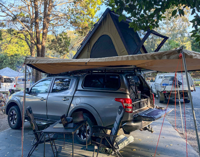 Here is a descriptive alt text for "roof tent camping set up woody head NSW.jpg":"A grey 4x4 ute parked at a wooded campsite, set up with an open hardshell rooftop tent and a deployed tan side awning. Underneath the awning, a temporary workspace features a small folding table with a laptop and two black camp chairs. The ute's tailgate is down, revealing a built-in drawer storage system inside the canopy.