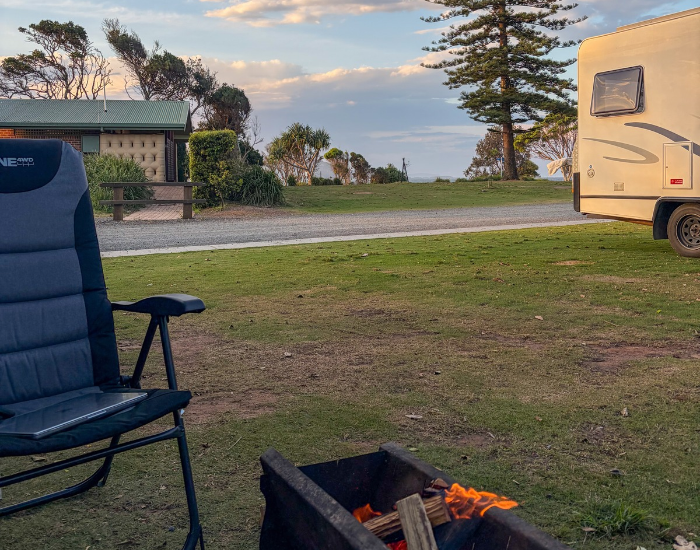 camping chair with a laptop set up next to a crackling fire pit at a grassy campground near amenities and a camper trailer.