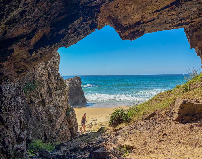 A person walking along a sandy beach as viewed through the opening of a rocky coastal cave looking out toward the ocean.