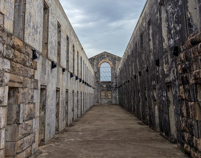 A central walkway through the historic, roofless stone cellblock of Trial Bay Gaol with stone walls and window openings leading toward a large arched window at the end